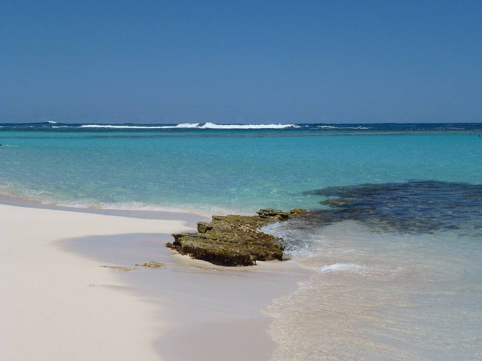 A rock ledge running into clear water at Loblolly Bay, with the reef dark under the surface and surf breaking on the outer reef