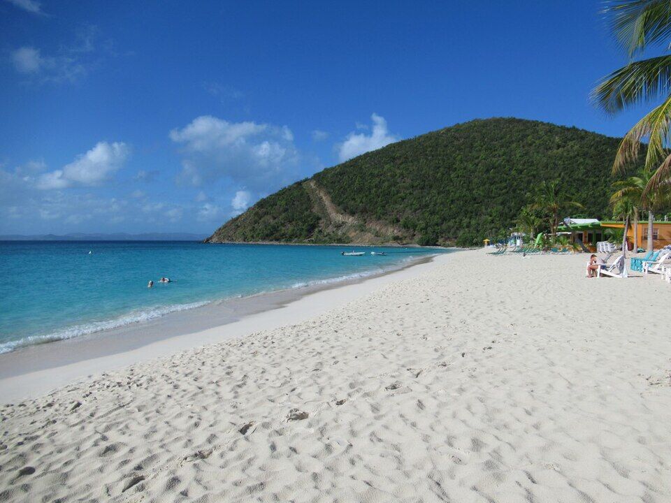 The main beach looking west to the hill at the end of the bay, with loungers and a beach bar under the palms (December 2019)