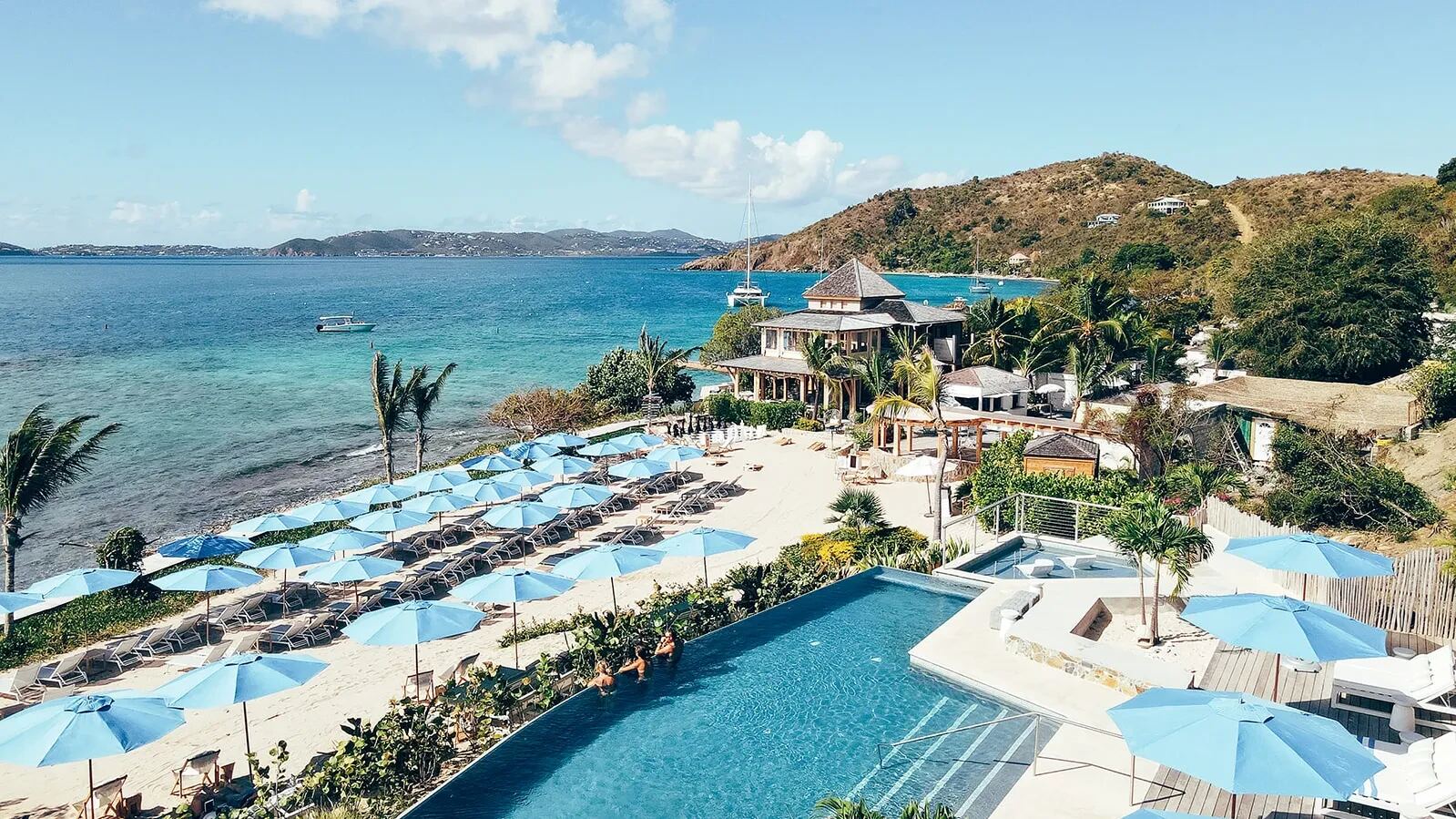 Lovango's Beach Club from above: the infinity pool, rows of blue umbrellas on the sand and the clubhouse, with yachts at anchor and the islands across Pillsbury Sound